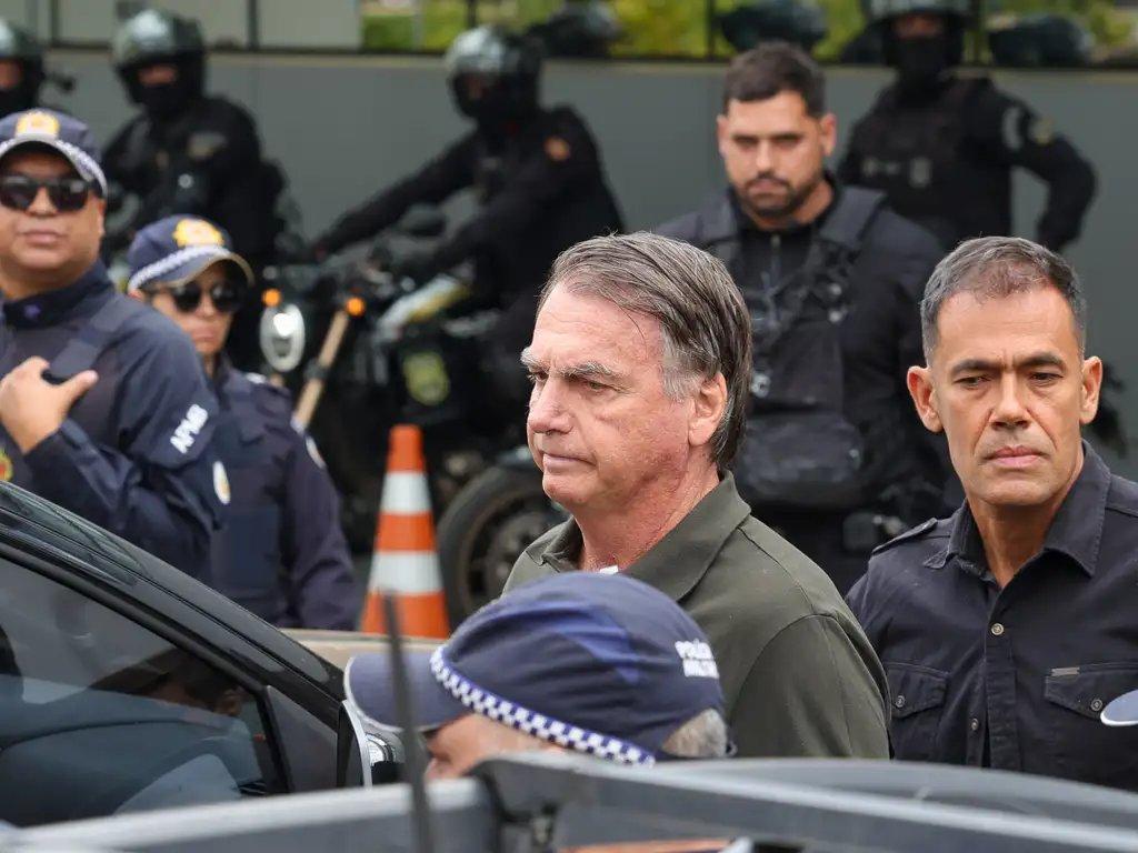 Bolsonaro é escoltado por agentes de segurança e policiais uniformizados em área externa, com motos e cones ao fundo.