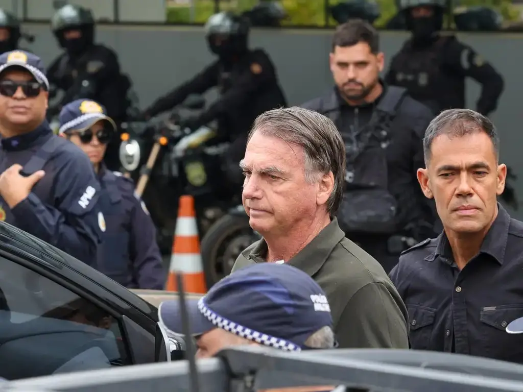 Bolsonaro é escoltado por agentes de segurança e policiais uniformizados em área externa, com motos e cones ao fundo.