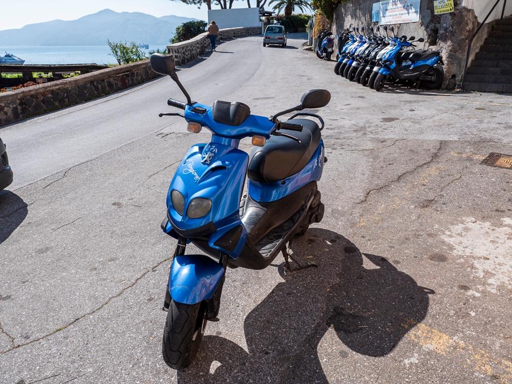 Na imagem, uma fotografia em ângulo médio e luz solar intensa mostra uma motoneta (scooter) azul brilhante estacionada em primeiro plano em uma rua pavimentada. A motoneta está levemente inclinada sobre o descanso lateral, com o guidão virado para a esquerda e o logotipo da Peugeot visível na parte frontal. Ao fundo, à direita, há uma fileira de várias outras motonetas azuis idênticas estacionadas contra um muro de pedra, sugerindo um ponto de aluguel. No topo do muro, há placas informativas e o nome 