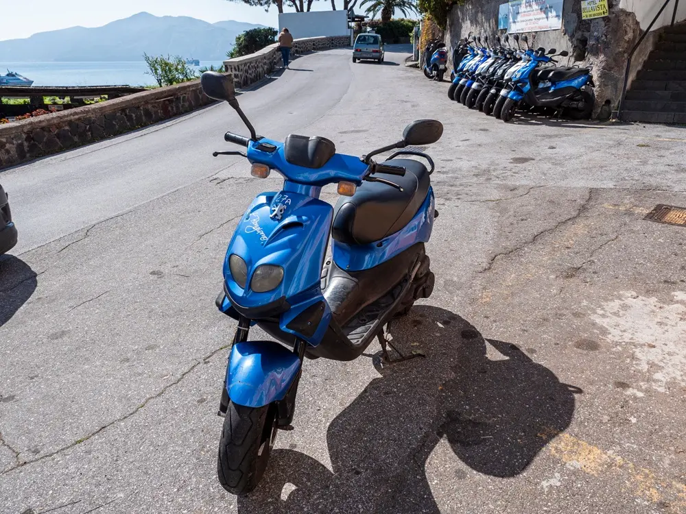 Na imagem, uma fotografia em ângulo médio e luz solar intensa mostra uma motoneta (scooter) azul brilhante estacionada em primeiro plano em uma rua pavimentada. A motoneta está levemente inclinada sobre o descanso lateral, com o guidão virado para a esquerda e o logotipo da Peugeot visível na parte frontal. Ao fundo, à direita, há uma fileira de várias outras motonetas azuis idênticas estacionadas contra um muro de pedra, sugerindo um ponto de aluguel. No topo do muro, há placas informativas e o nome 