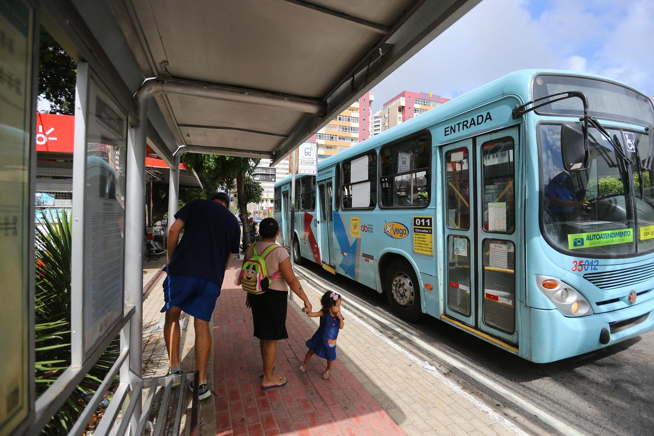 Passageiros aguardam sob o abrigo de um ponto de ônibus em Fortaleza, enquanto um coletivo azul claro estaciona para embarque. Uma mulher segurando a mão de uma criança pequena em destaque no primeiro plano na parada de ônibus.