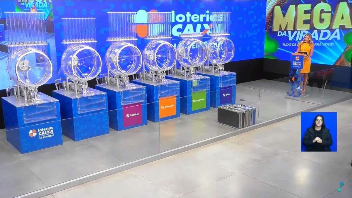 Esta imagem captura o estúdio de um sorteio das Loterias CAIXA, especificamente para a Mega da Virada. No centro, seis globos de acrílico transparente estão alinhados sobre bases azuis; cada base exibe o logotipo de um jogo diferente, como Lotofácil e Quina, em cores vibrantes como rosa, laranja e verde. À direita, uma apresentadora loira, vestindo um vestido amarelo, está posicionada atrás de um púlpito transparente, enquanto o fundo é composto por grandes painéis digitais azuis com o nome do sorteio. No canto inferior direito, há um pequeno quadro em destaque mostrando uma intérprete de LIBRAS realizando a tradução em tempo real. O ambiente é amplamente iluminado, com um piso cinza claro que reflete as luzes e a estrutura do estúdio, transmitindo uma atmosfera profissional e oficial.