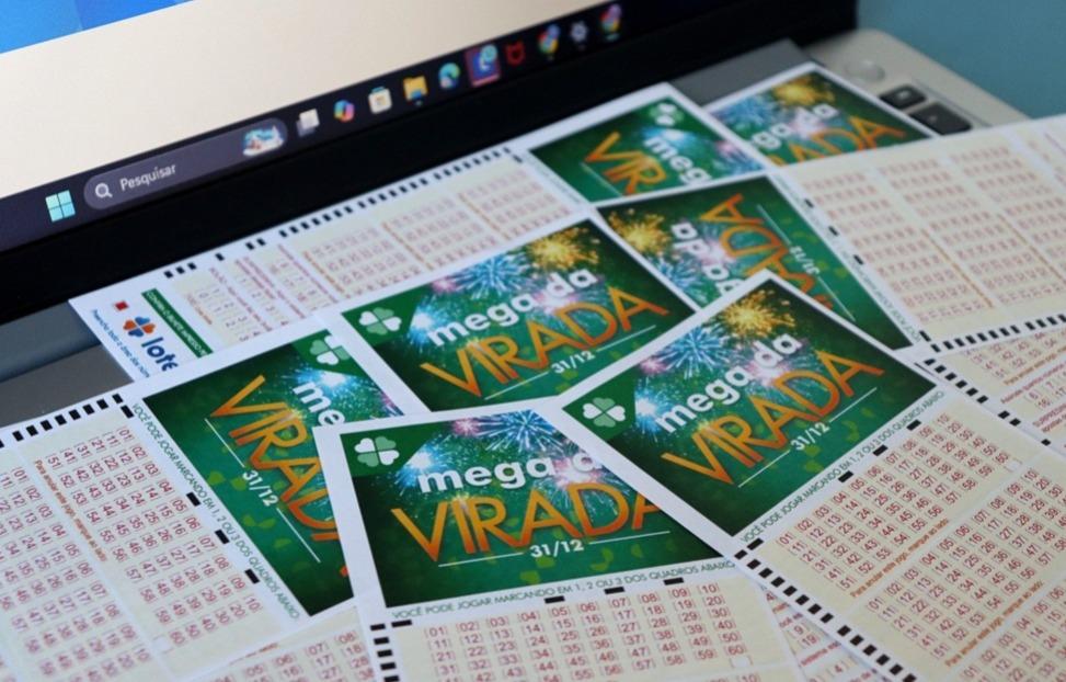 Apostas da Mega da Virada em cima de mesa próximo a notebook.