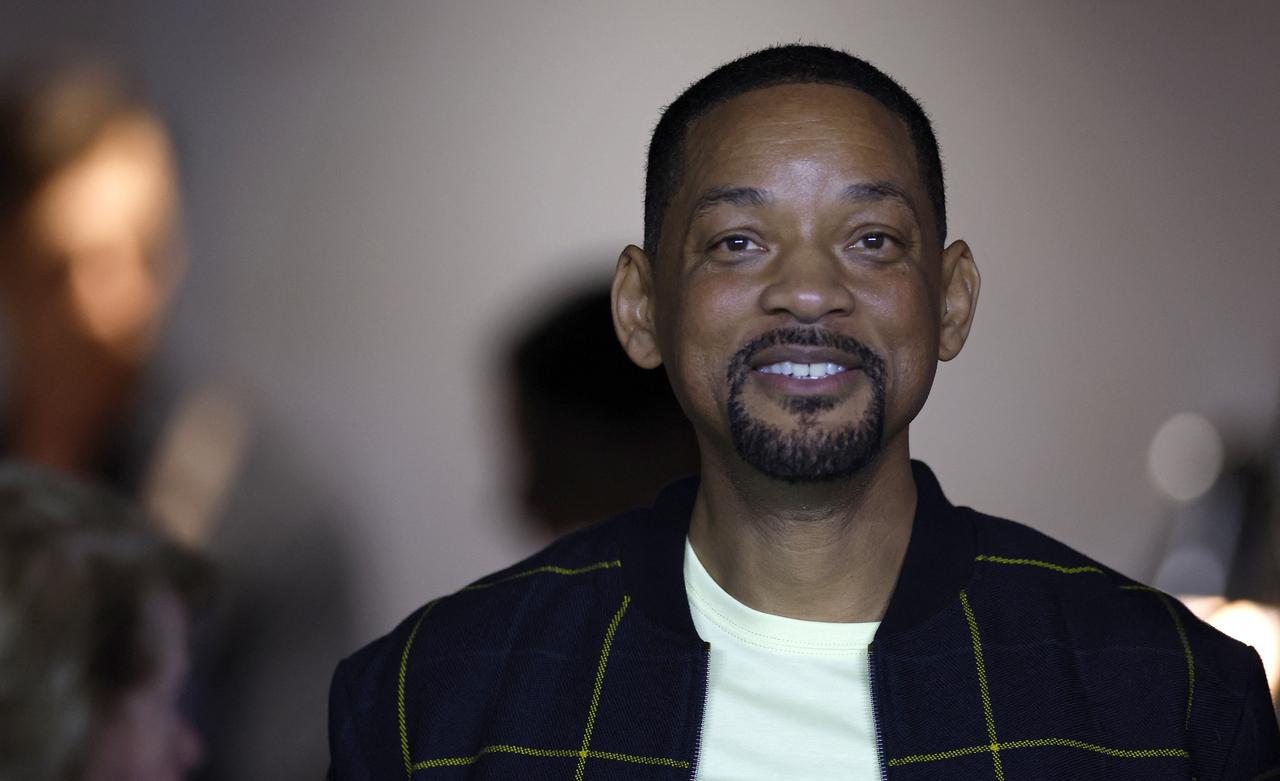 Foto mostra ator Will Smith de camiseta branca, com jaqueta preta, sorrindo.