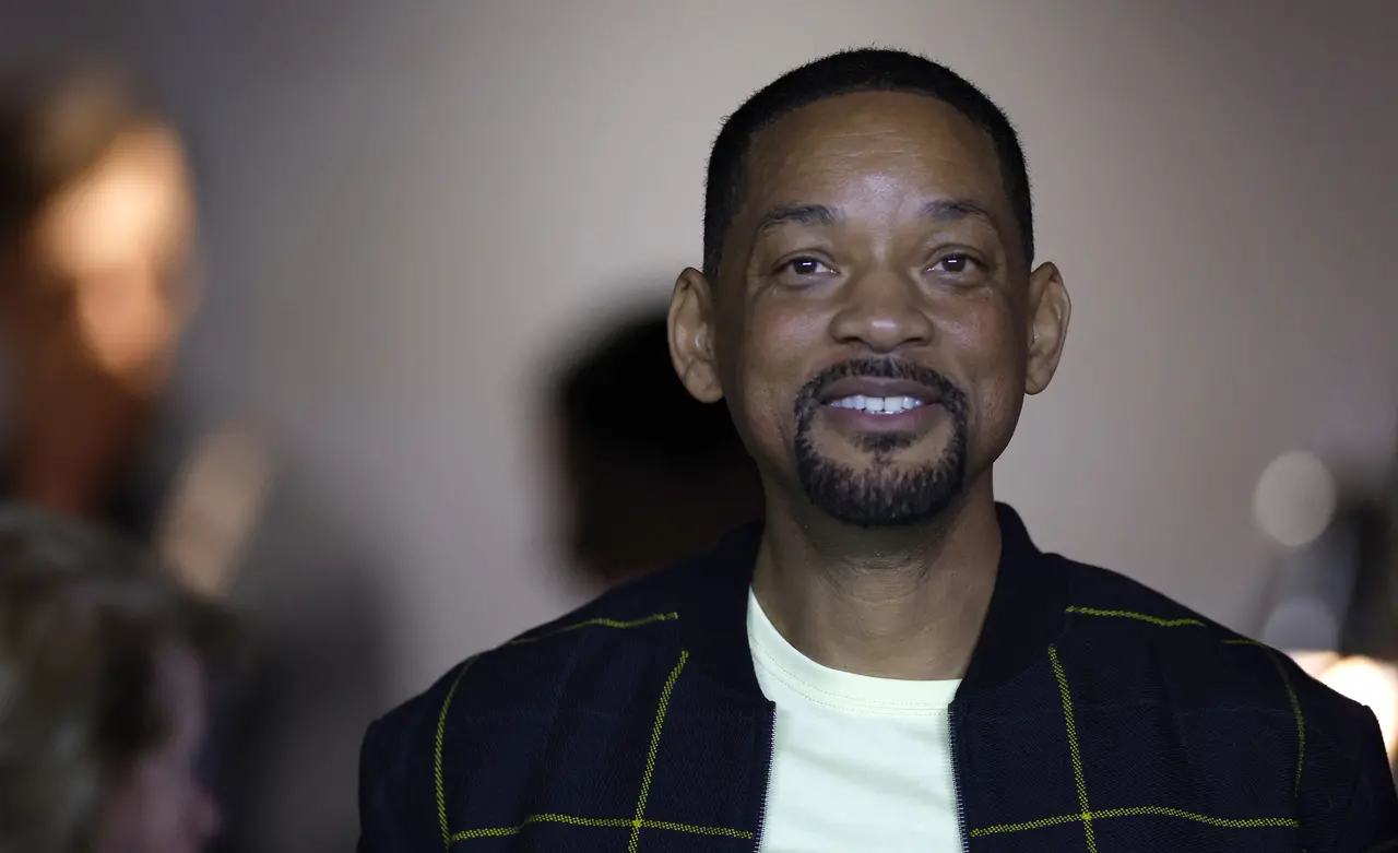 Foto mostra ator Will Smith de camiseta branca, com jaqueta preta, sorrindo.