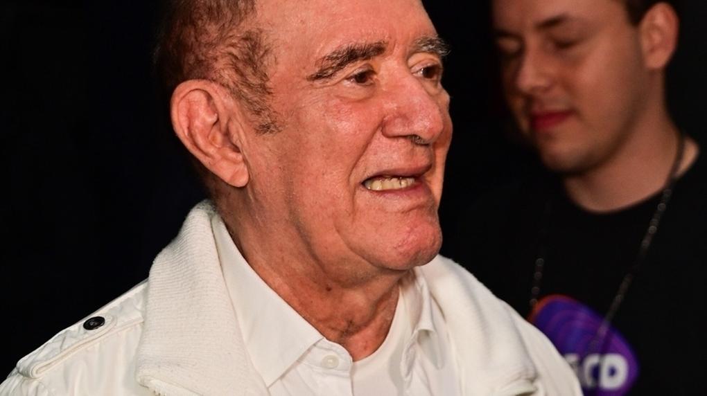 Esta imagem apresenta um retrato de meio-corpo do renomado humorista brasileiro Renato Aragão, conhecido como Didi. Ele é um homem branco, de idade avançada, com rugas expressivas ao redor dos olhos e da boca, e cabelos curtos e grisalhos. Ele está posicionado de perfil parcial, voltado para a direita, exibindo um sorriso suave que revela os dentes superiores. Veste uma jaqueta branca de estilo esportivo com zíperes e botões, sobreposta a uma camisa social também branca que possui uma pequena estrela escura bordada no peito. O fundo é escuro e levemente desfocado, onde se nota a presença parcial de outra pessoa à direita e uma mão com unhas vermelhas em primeiro plano, sugerindo que ele está em um evento social ou sendo entrevistado.