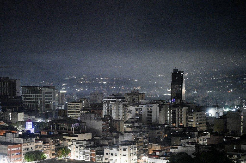 Na imagem, vista panorâmica noturna da cidade de Caracas sob um céu nublado e escuro. Em primeiro plano, diversos edifícios residenciais e comerciais de diferentes alturas estão iluminados por luzes internas e públicas. À direita, destaca-se um edifício mais alto e escuro que sobressai no horizonte. Ao fundo, as luzes da cidade estendem-se pelas encostas das montanhas, parcialmente cobertas por uma névoa baixa e densa. A iluminação geral cria um contraste forte entre o brilho das janelas e as sombras profundas da noite.