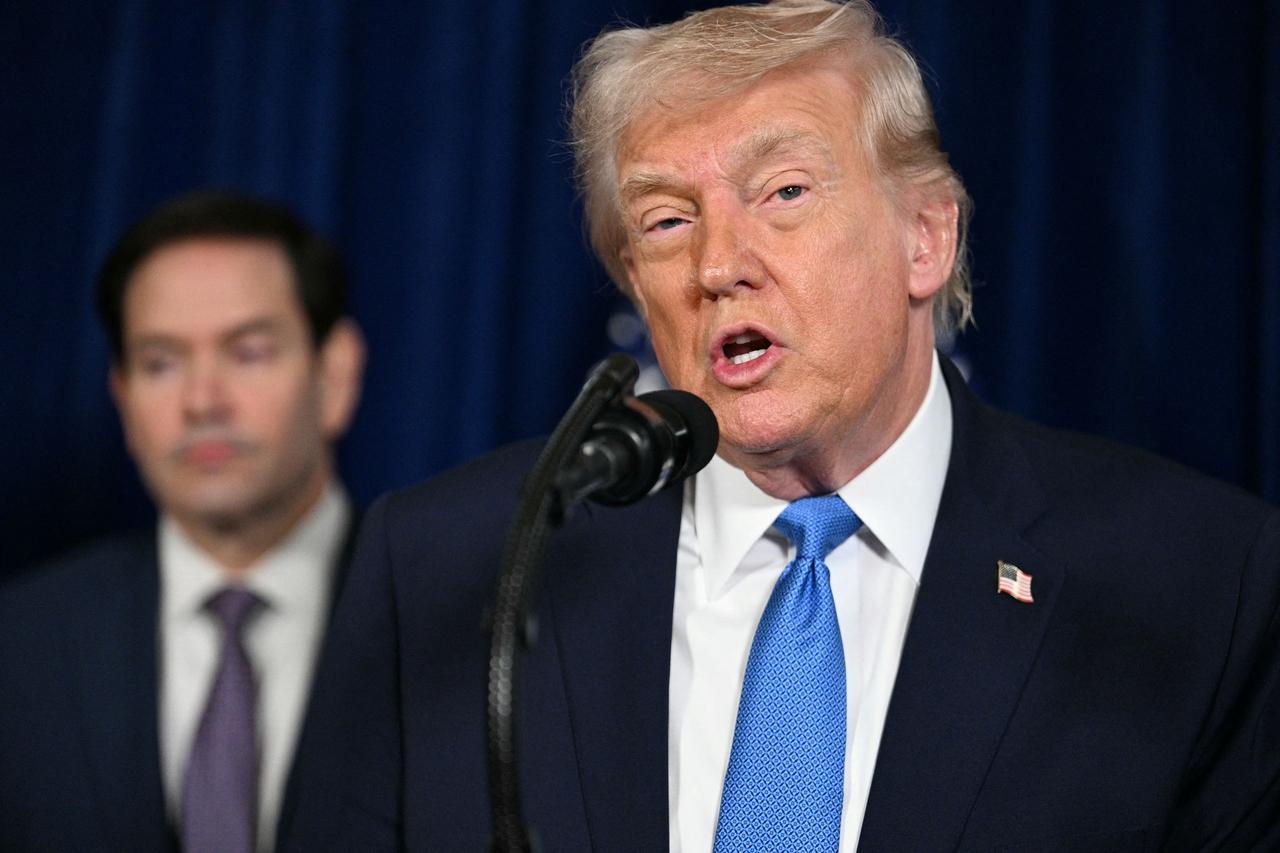 Donald Trump discursa cem frente a um microfone, vestindo terno azul escuro e gravata azul clara sobre um fundo de cortina azul. Ao fundo, Marco Rubio está em desfoque.