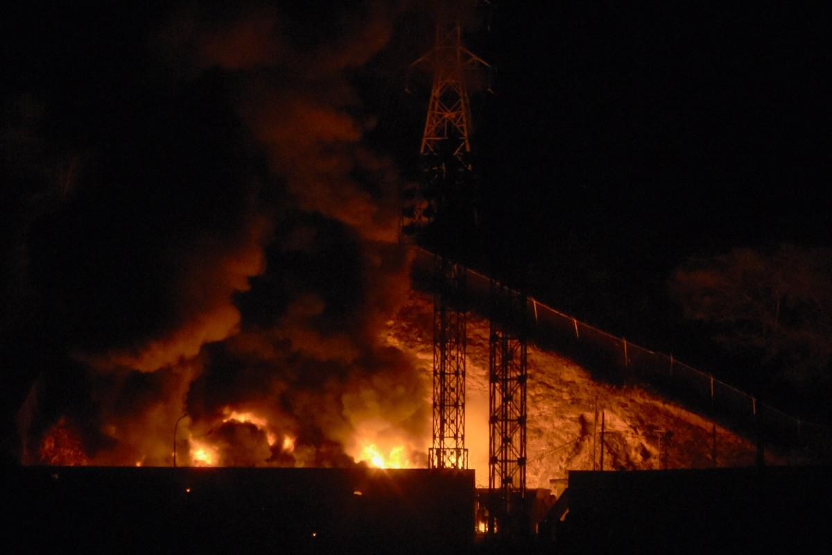 Explosões e incêndio de grandes proporções iluminam a noite, com chamas e fumaça densa ao fundo de estruturas metálicas e torres industriais.