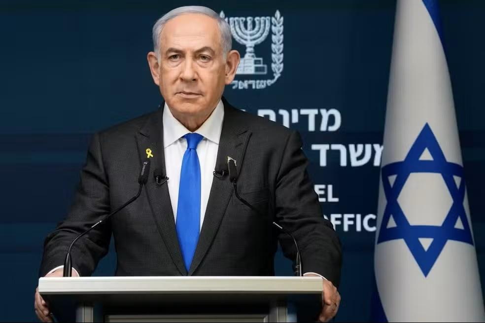 Na imagem, fotografia em plano médio do Primeiro-Ministro de Israel, Benjamin Netanyahu, falando atrás de um púlpito cinza. Ele veste um terno preto, camisa branca e gravata azul vibrante. Na lapela direita, usa um pequeno broche de fita amarela. Ao fundo, à esquerda, vê-se o brasão de Israel em uma parede azul escura com dizeres em hebraico e inglês; à direita, parte da bandeira de Israel com a Estrela de Davi azul.