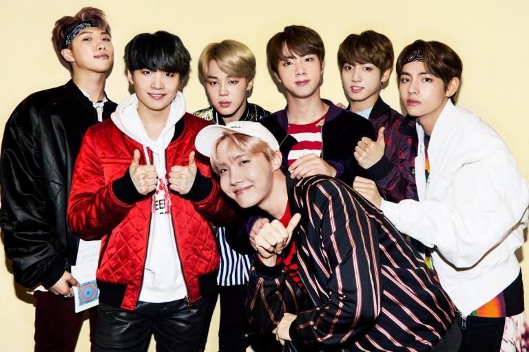 Na imagem, fotografia colorida do grupo de K-pop BTS, com os sete integrantes posando juntos contra um fundo amarelo claro. Eles vestem jaquetas de estilo casual e streetwear.