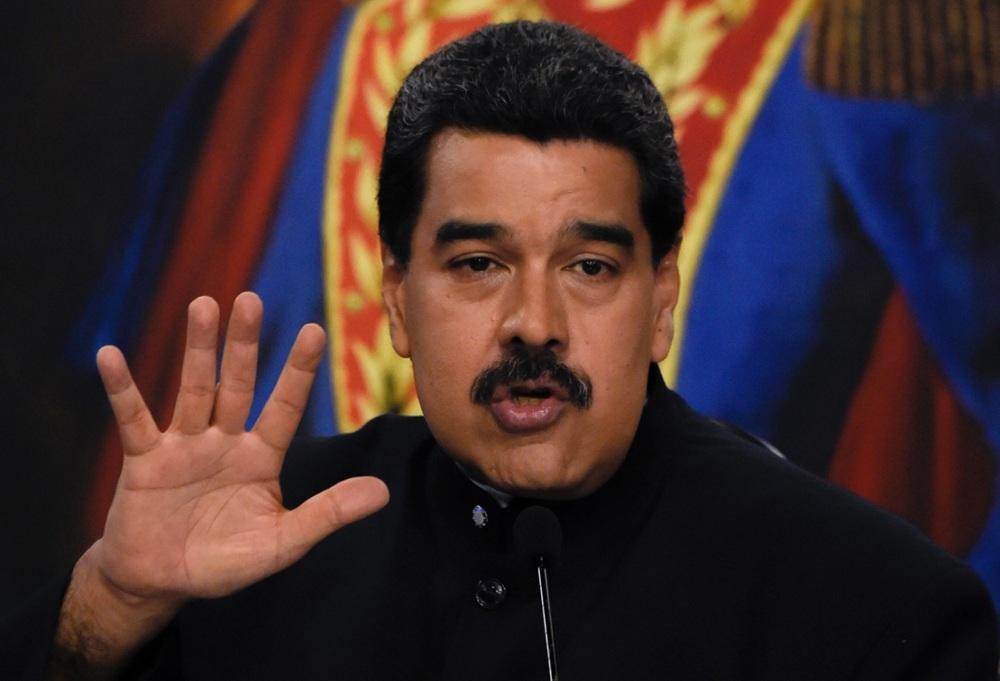 Na imagem, o presidente da Venezuela, Nicolás Maduro, é retratado do peito para cima contra um fundo desfocado que sugere uma bandeira ou estandarte oficial. Ele tem cabelos escuros, bigode farto e está com a boca levemente aberta como se estivesse discursando. Sua mão direita está erguida à frente, com os dedos estendidos e a palma voltada para a câmera. Ele veste uma túnica preta de gola alta com botões prateados. Um microfone preto está posicionado logo abaixo de seu rosto.
