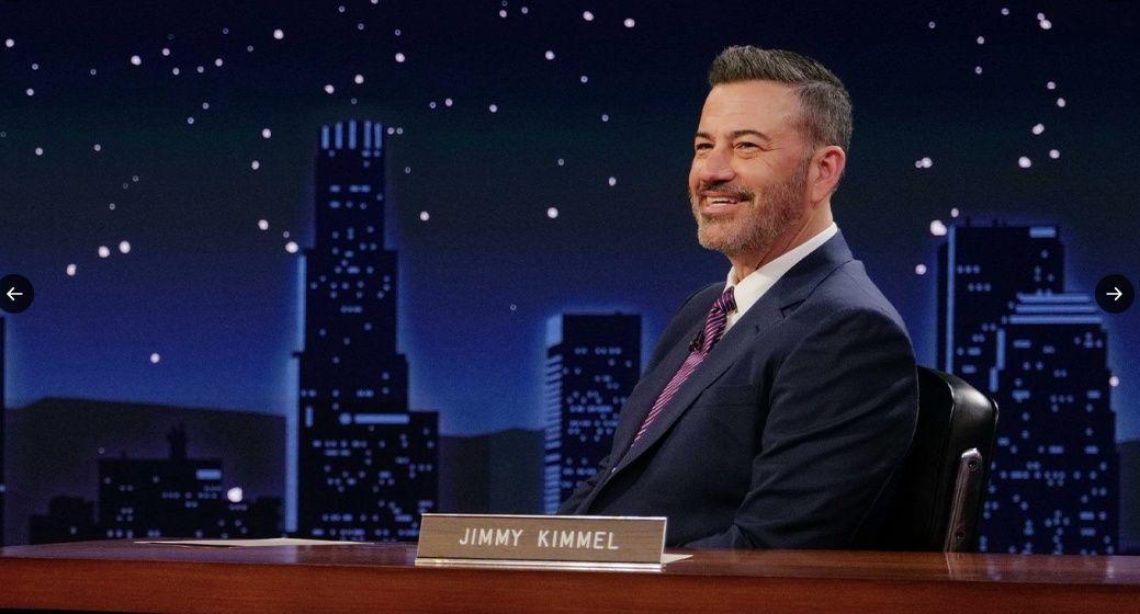 Jimmy Kimmel, o apresentador de talk show, sorrindo e sentado em sua mesa, com um cenário noturno de arranha-céus ao fundo.
