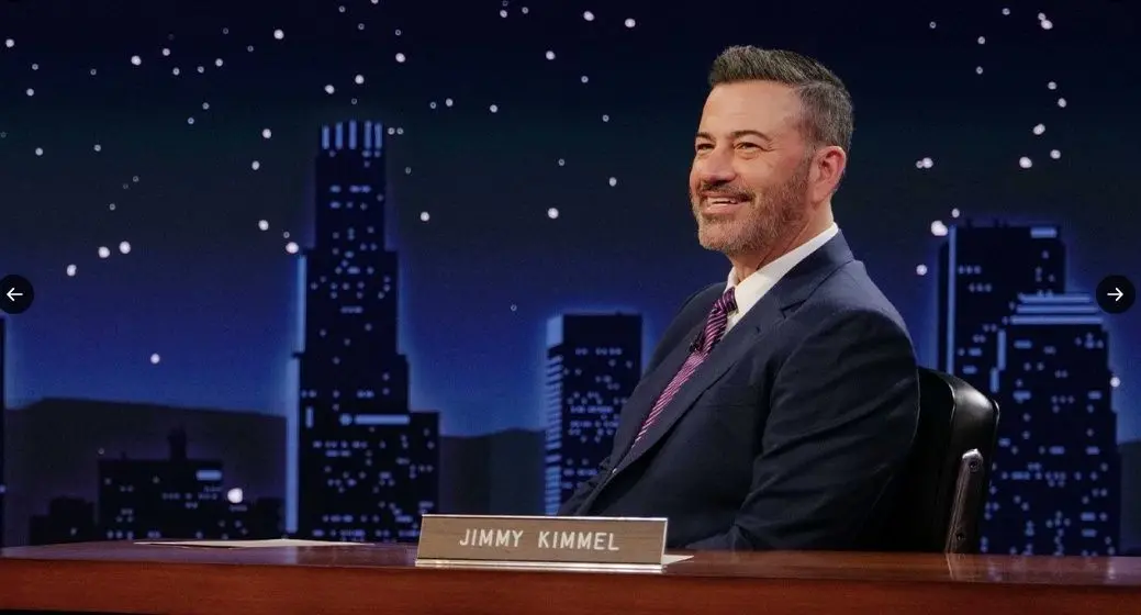 Jimmy Kimmel, o apresentador de talk show, sorrindo e sentado em sua mesa, com um cenário noturno de arranha-céus ao fundo.