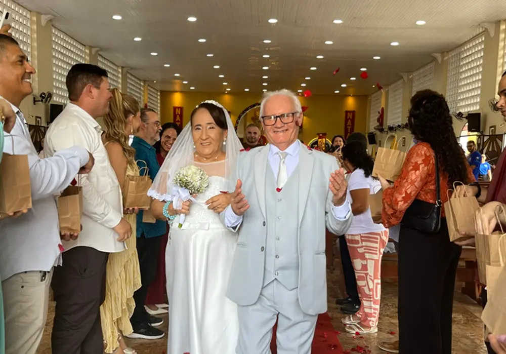 Na imagem, uma fotografia colorida de um casal de idosos, Edilton e Francisca, caminhando radiantes pelo corredor central de uma igreja após a cerimônia de casamento. A noiva, à esquerda, usa um vestido branco com detalhes em renda, véu e segura um buquê de flores brancas. O noivo, à direita, veste um terno cinza claro com colete e gravata prateada, usa óculos de armação escura e sorri com os braços levemente abertos. Eles caminham sobre um tapete vermelho enquanto convidados, posicionados em ambos os lados, sorriem e seguram sacolas de papel pardo (provavelmente contendo pétalas ou lembranças). Pétalas de rosas vermelhas são vistas caindo no ar ao redor do casal. O interior da igreja possui teto de madeira clara, iluminação embutida e paredes com detalhes vazados.