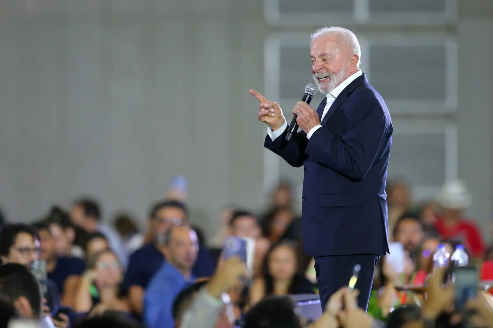 Lula fala ao microfone durante evento com público, veste terno escuro e aponta com o dedo enquanto sorri; pessoas ao fundo acompanham e registram com celulares