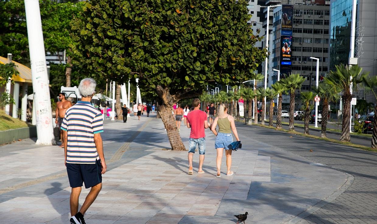 Pessoas caminham pelo calçadão ensolarado da Beira-Mar, em Fortaleza, durante uma onda de calor. A cena mostra pedestres em trajes leves sob a sombra de árvores, com prédios altos e palmeiras ao fundo sob luz solar intensa.