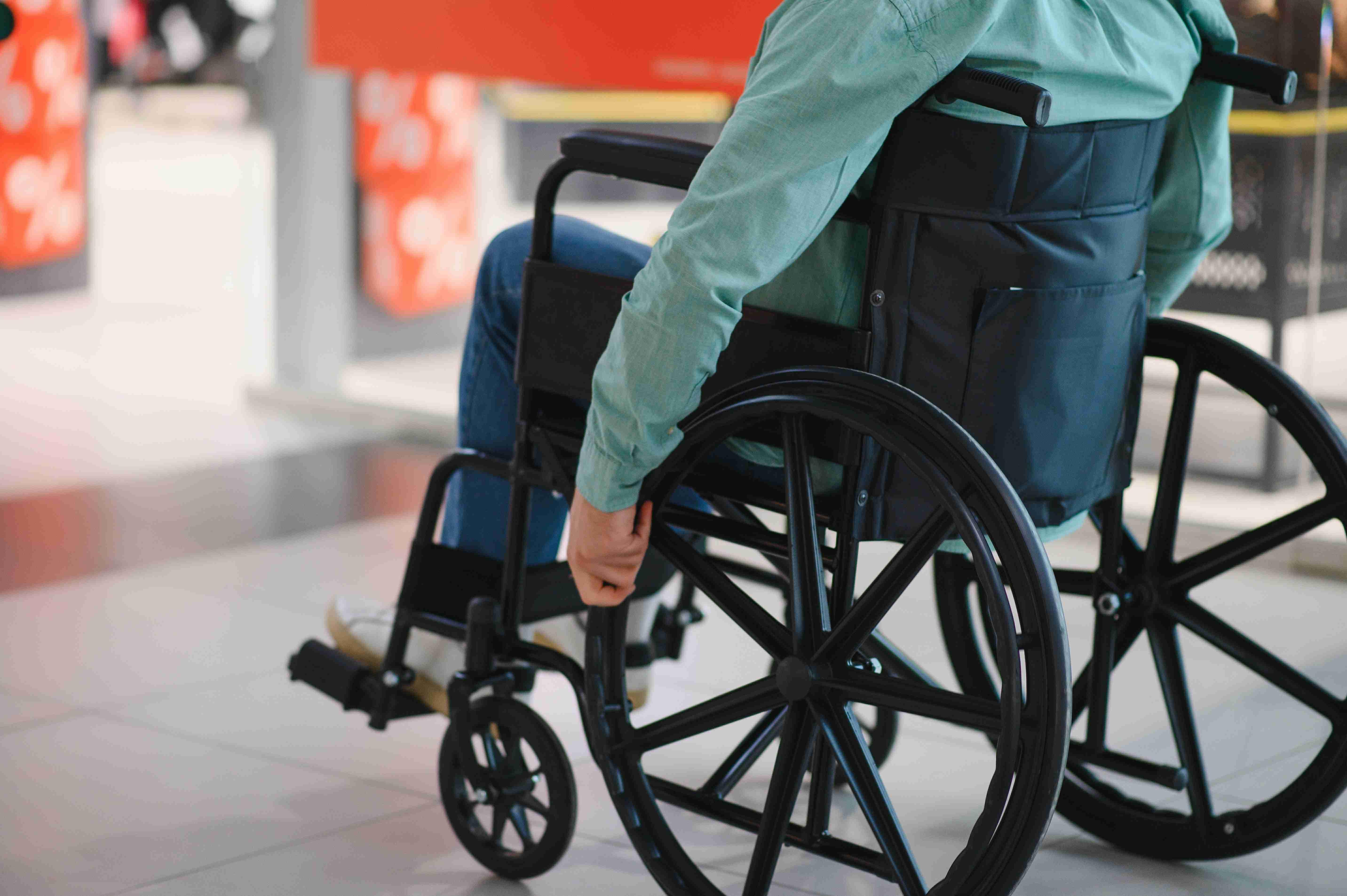 Homem em cadeira de rodas, vestido com camisa verde e jeans, impulsionando-se em um espaço público.