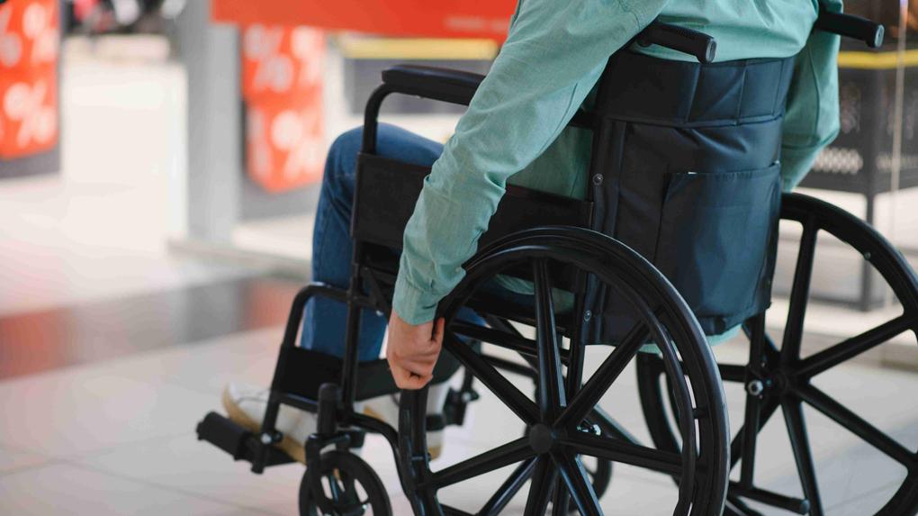 Homem em cadeira de rodas, vestido com camisa verde e jeans, impulsionando-se em um espaço público.
