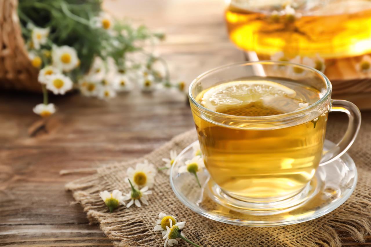 Xícara de chá de camomila quente com uma rodela de limão, colocada sobre um tecido rústico, com pequenas flores de camomila espalhadas ao redor e um bule ao fundo em uma mesa de madeira.