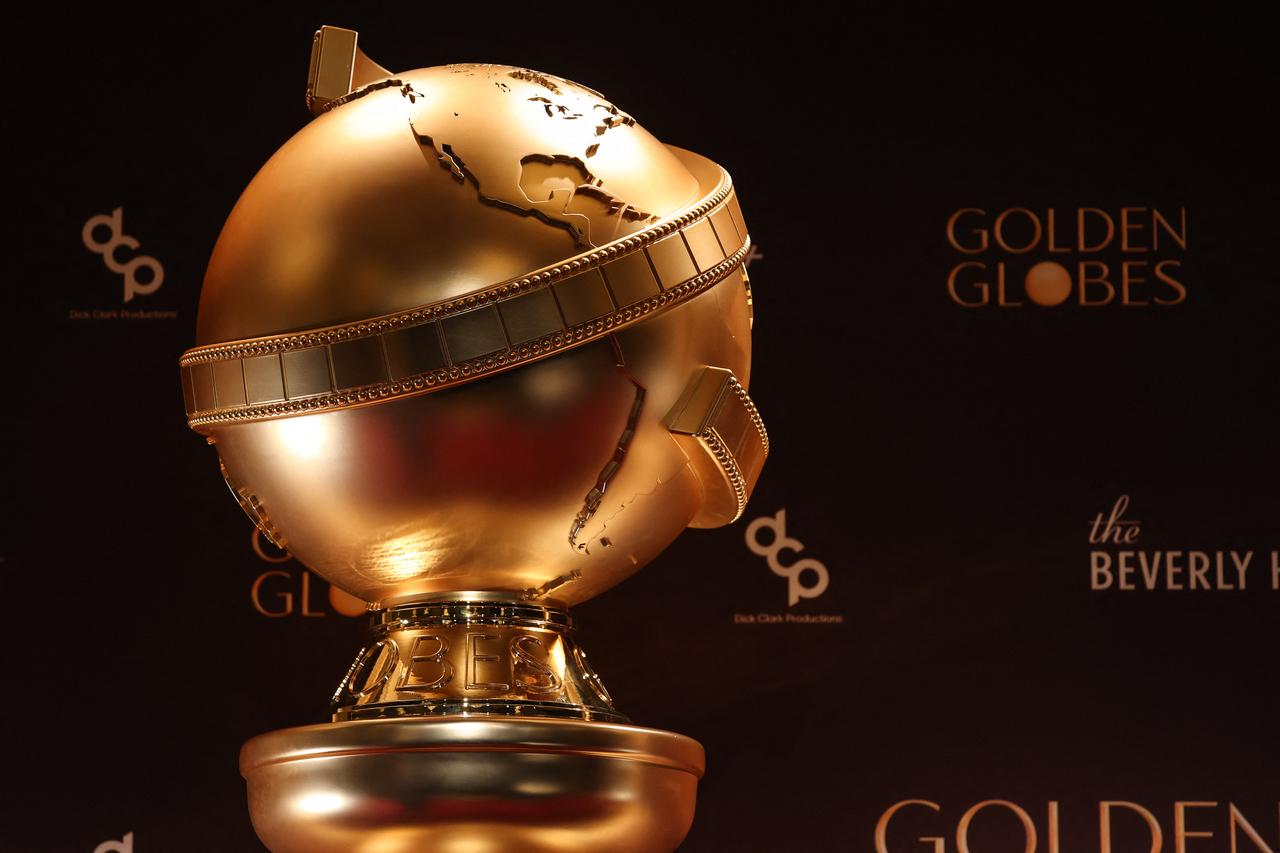 O troféu dourado do Golden Globes aparece em destaque, exibindo o globo terrestre envolto por uma fita de película cinematográfica sobre uma base metálica. O fundo é composto por uma parede preta com os logotipos da premiação e da Dick Clark Productions aplicados em cor dourada.