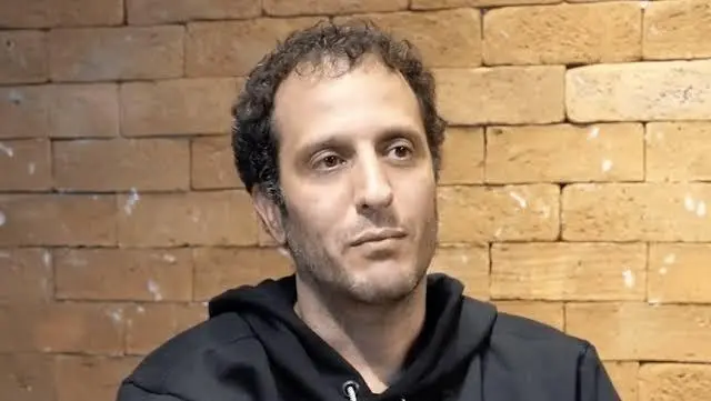 A foto mostra um homem branco, calvo e barba rasa, vestido com uma blusa preta, à frente de uma parede marrom.