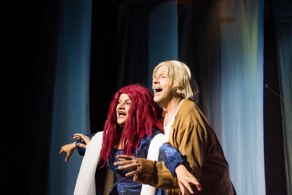 Na imagem, dois atores em cena no palco, recriando a icônica pose do filme Titanic. À frente, uma mulher com uma peruca volumosa de fios vermelhos intensos e um vestido azul escuro, com os braços abertos e uma expressão de empolgação. Atrás dela, um homem com peruca loira e casaco marrom a segura pela cintura, também com a boca aberta em um semblante alegre ou de canto. O fundo é composto por cortinas em tons de azul e iluminação cênica focada nos personagens.