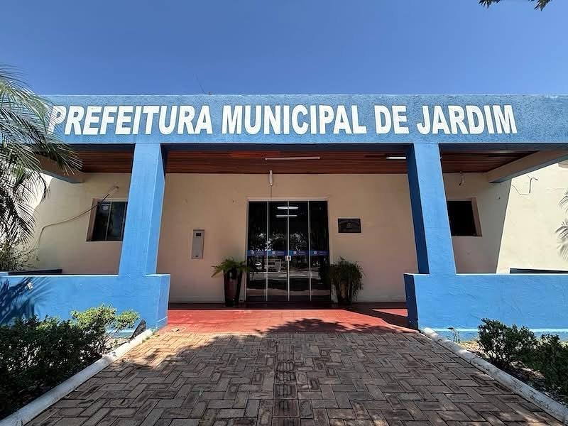 Fachada principal da Prefeitura Municipal de Jardim, com a inscrição 'PREFEITURA MUNICIPAL DE JARDIM' em destaque na cor azul, sob um céu claro. O edifício apresenta colunas e detalhes em azul, paredes claras e uma entrada de vidro, com um caminho de tijolos que leva até a porta.