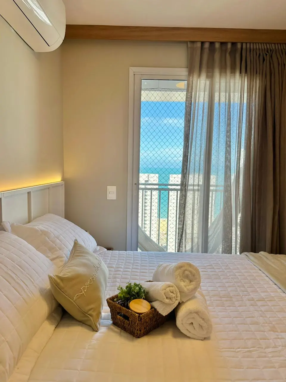 Na imagem, um quarto de casal com decoração clean e vista para o mar. A cama está arrumada com um edredom branco texturizado, travesseiros coordenados e uma cesta de palha com toalhas brancas enroladas e um pequeno vaso de planta. Ao fundo, uma porta de vidro com rede de proteção dá acesso a uma varanda, revelando prédios litorâneos e o azul do oceano sob um céu claro. A iluminação é suave, com um detalhe de fita LED embutida na cabeceira branca e cortinas translúcidas em tom de fendi.