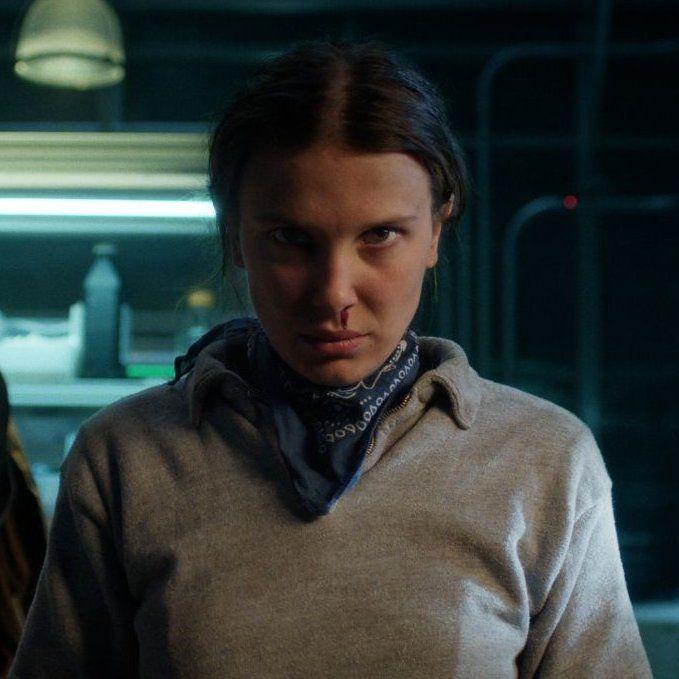 Millie Bobby Brown (como Eleven) com expressão séria e determinada, olhando diretamente para a câmera, em um cenário escuro com iluminação artificial.