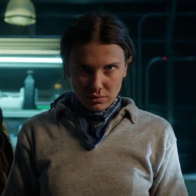 Millie Bobby Brown (como Eleven) com expressão séria e determinada, olhando diretamente para a câmera, em um cenário escuro com iluminação artificial.