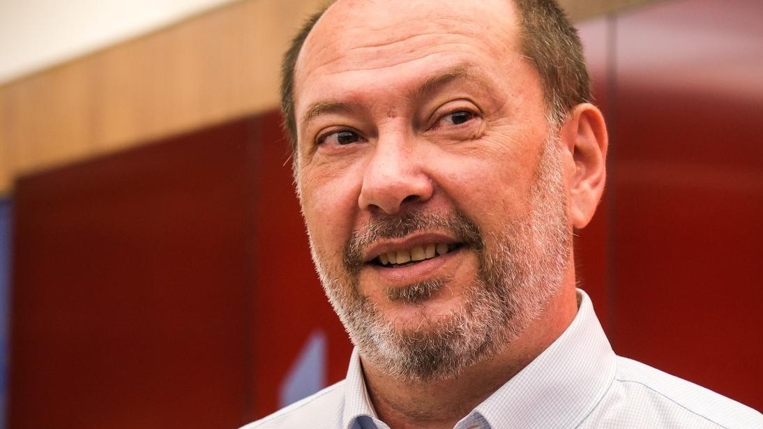 Retrato em close de Ivo Gomes, com entradas no cabelo e barba curta grisalha, usando camisa clara. Ele sorri levemente e olha para fora da câmera. Ao fundo, há um painel em tom vermelho.