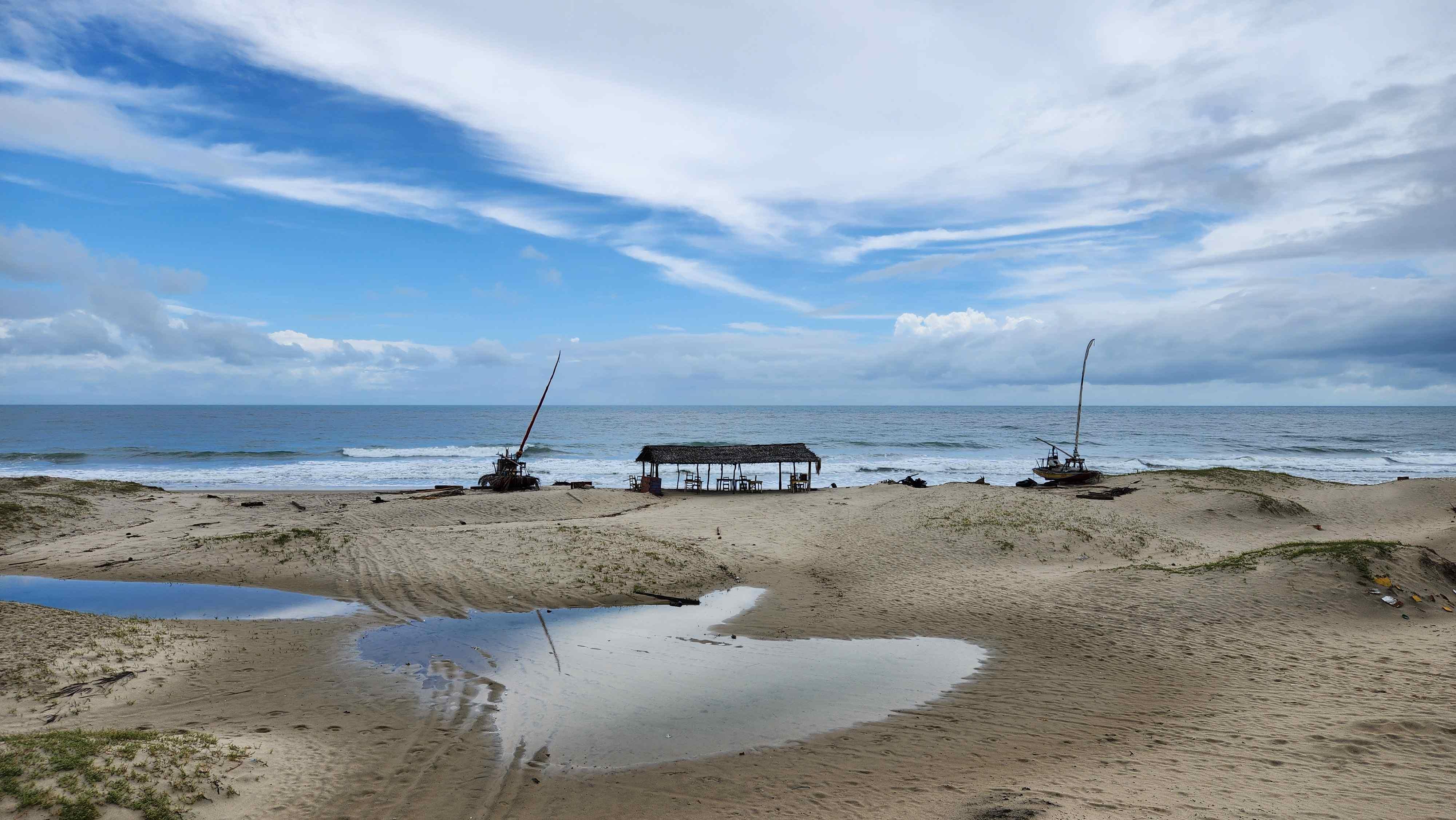 Vista panorâmica da Praia do Balbino em Cascavel, Ceará, sob um céu azul com nuvens esparsas. Em primeiro plano, uma faixa de areia clara abriga uma barraca rústica de madeira com teto de palha, onde se veem mesas e cadeiras. À esquerda, uma pequena embarcação do tipo jangada está estacionada na areia, enquanto o mar calmo e o horizonte completam o cenário ao fundo.