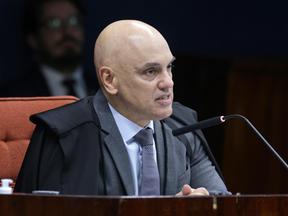 Autoridade judicial sentada à bancada durante uma sessão formal, vestindo toga sobre terno e gravata, falando ao microfone em um ambiente institucional com mesa de madeira e cadeira estofada ao fundo, em contexto de julgamento ou audiência. A autoridade em questão é Alexandre de Moraes.