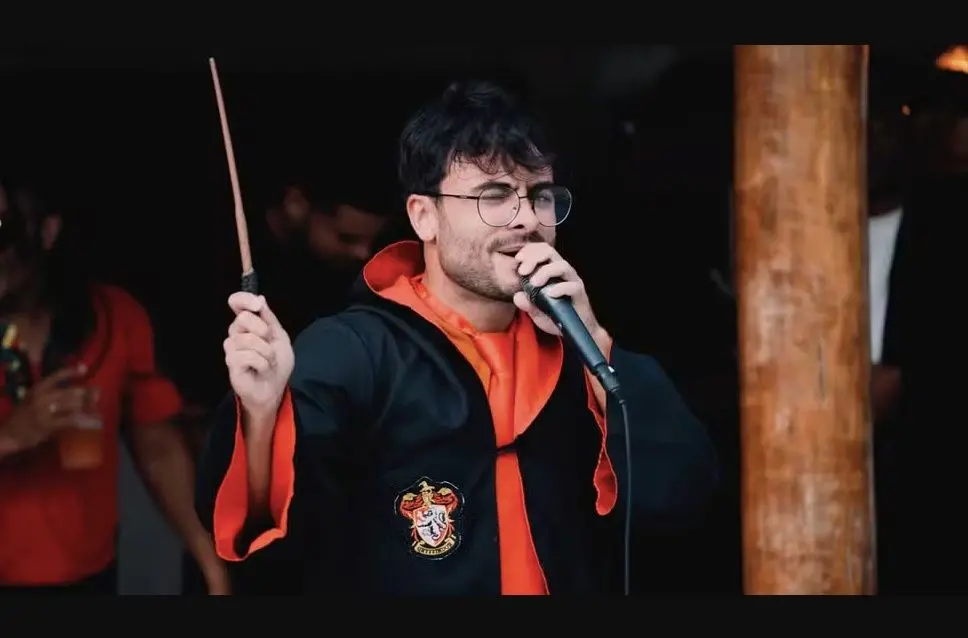 Homem vestido como Harry Potter, com óculos, segurando uma varinha mágica e cantando em um microfone.