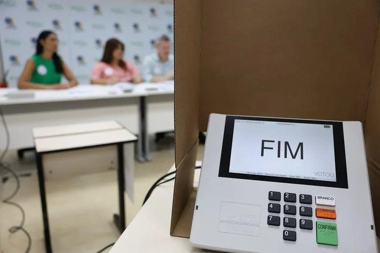 Urna eletrônica em primeiro plano, com a tela exibindo a palavra ‘FIM’ após o encerramento do voto. Ao fundo, desfocados, mesários sentados atrás de uma mesa em uma seção eleitoral.