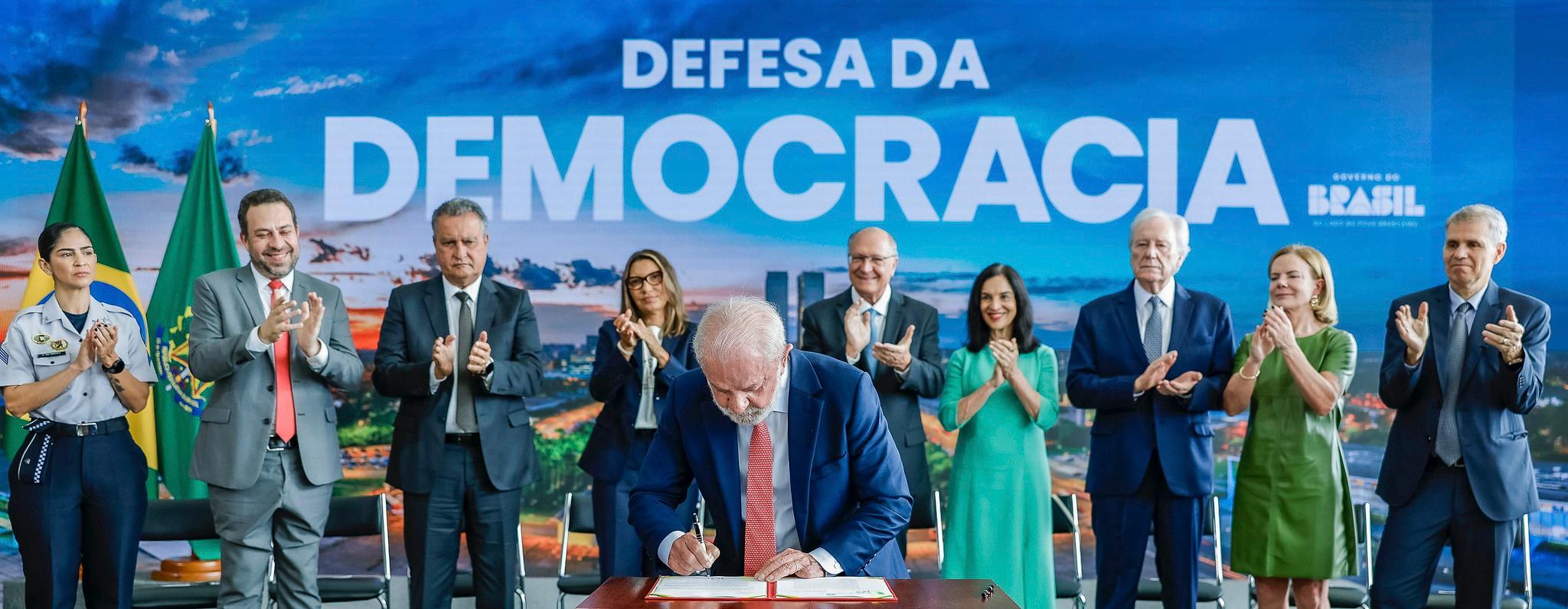 Autoridades em trajes formais estão reunidas em um palco institucional enquanto um representante assina um documento sobre uma mesa. Ao fundo, um grande painel azul exibe a frase 'Defesa da Democracia' e a marca do Governo do Brasil. Bandeiras nacionais aparecem à esquerda, e várias pessoas aplaudem o ato, sugerindo uma cerimônia solene de caráter oficial.