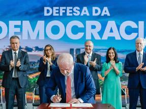 Autoridades em trajes formais estão reunidas em um palco institucional enquanto um representante assina um documento sobre uma mesa. Ao fundo, um grande painel azul exibe a frase 'Defesa da Democracia' e a marca do Governo do Brasil. Bandeiras nacionais aparecem à esquerda, e várias pessoas aplaudem o ato, sugerindo uma cerimônia solene de caráter oficial.