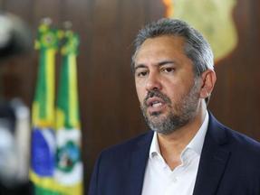 Elmano de Freitas de terno e camisa branca fala durante entrevista, com expressão séria, em ambiente institucional. Ao fundo, bandeiras do Brasil e do Ceará desfocadas; em primeiro plano, câmera de gravação.
