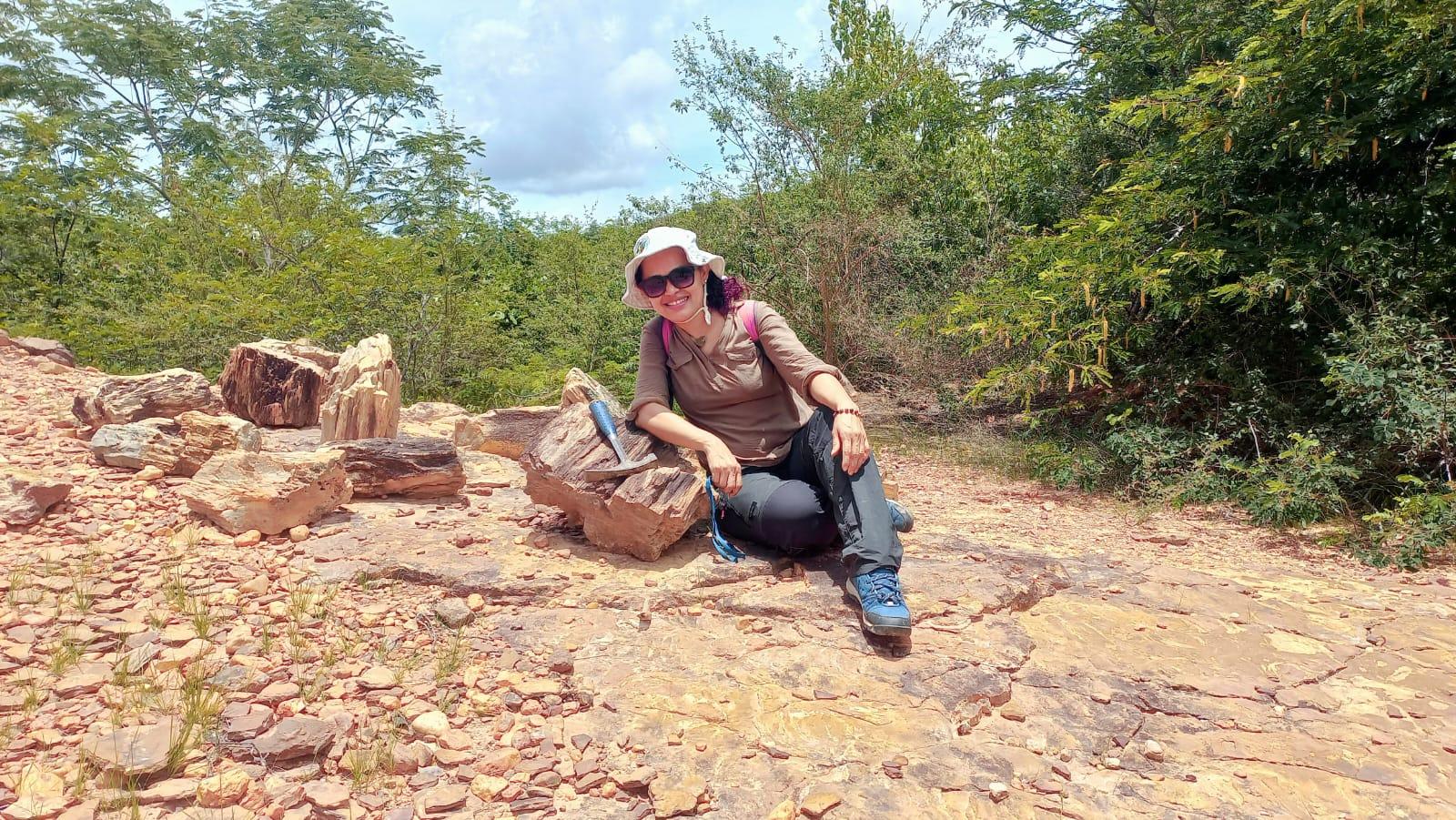 Mulher sorridente sentada em rochas, usando chapéu e óculos de sol, em paisagem com vegetação densa ao fundo. É Domingas Maria Conceição, pesquisadora.