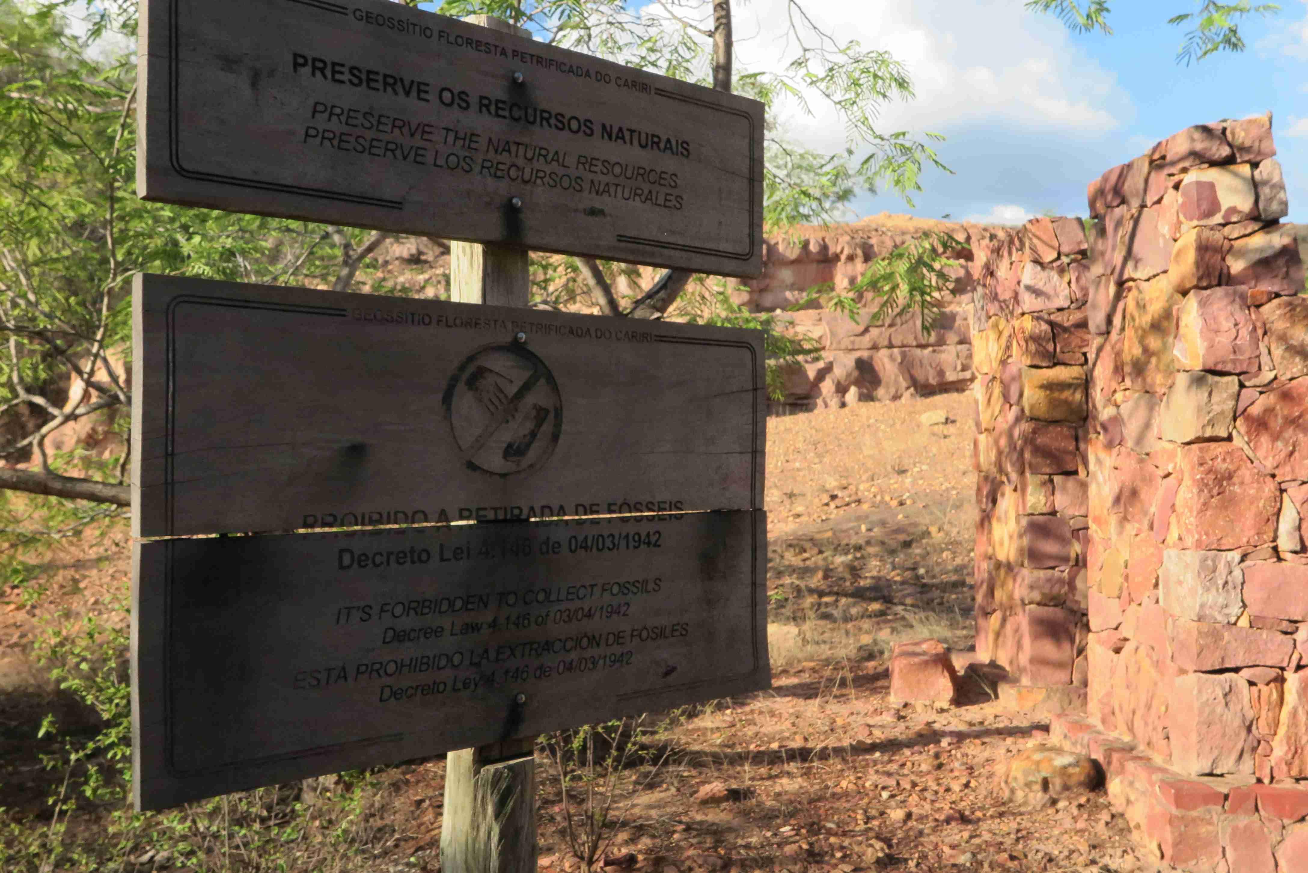 Duas placas de madeira informativas em frente a ruínas de pedra avermelhada em uma paisagem seca e rochosa, no Geopark Araripe, no Cariri cearense.