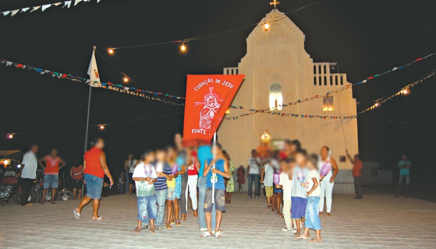 Celebração religiosa noturna em frente a uma igreja em Itatira. Um garoto segura um estandarte vermelho do Coração de Jesus em uma praça decorada com varais de luzes e bandeirinhas, rodeado por fiéis reunidos para a ocasião.