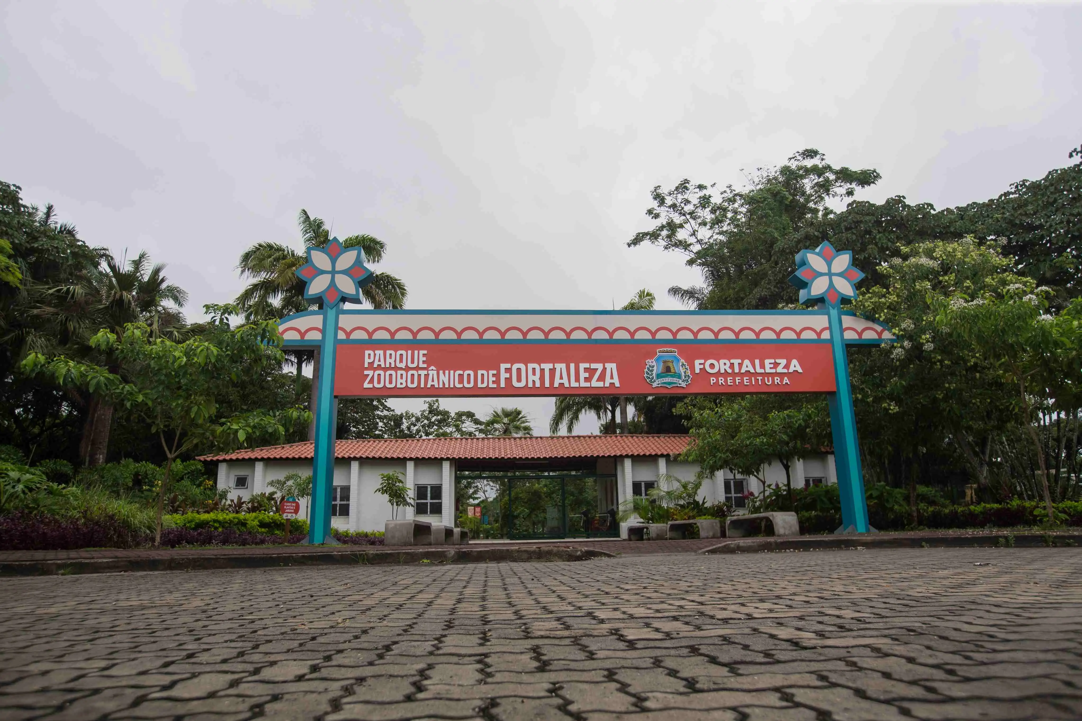 Fachada da entrada principal do Parque Zoobotânico de Fortaleza em um dia de céu encoberto. A imagem destaca um grande pórtico com detalhes em azul e vermelho, exibindo o letreiro com o nome do parque e o brasão da Prefeitura de Fortaleza.