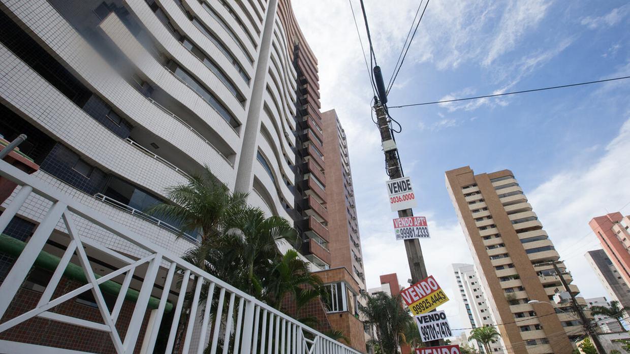 Foto que contém fachada de prédio de apartamentos com várias placas de venda.