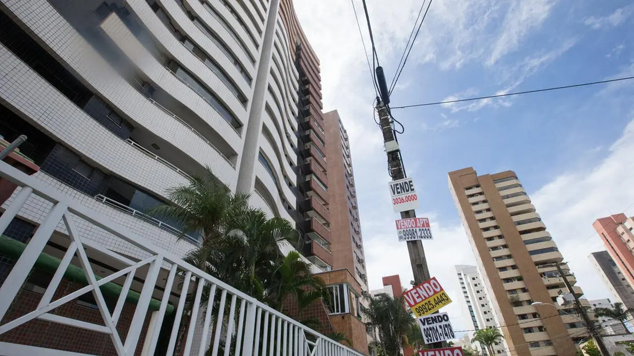 Foto que contém fachada de prédio de apartamentos com várias placas de venda.