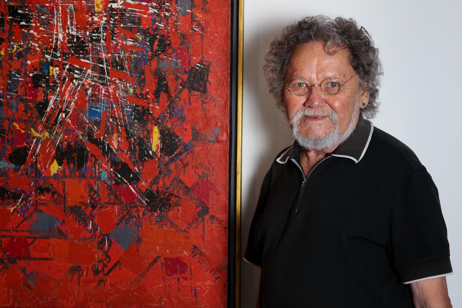 O artista visual cearense Zé Tarcísio posa ao lado de uma de suas obras, vestindo uma camisa preta e óculos redondos sob seus cabelos grisalhos e cacheados. A tela ao fundo apresenta uma composição abstrata vibrante com predominância de tons de vermelho, preto e azul, criando um contraste intenso com o fundo branco da parede.