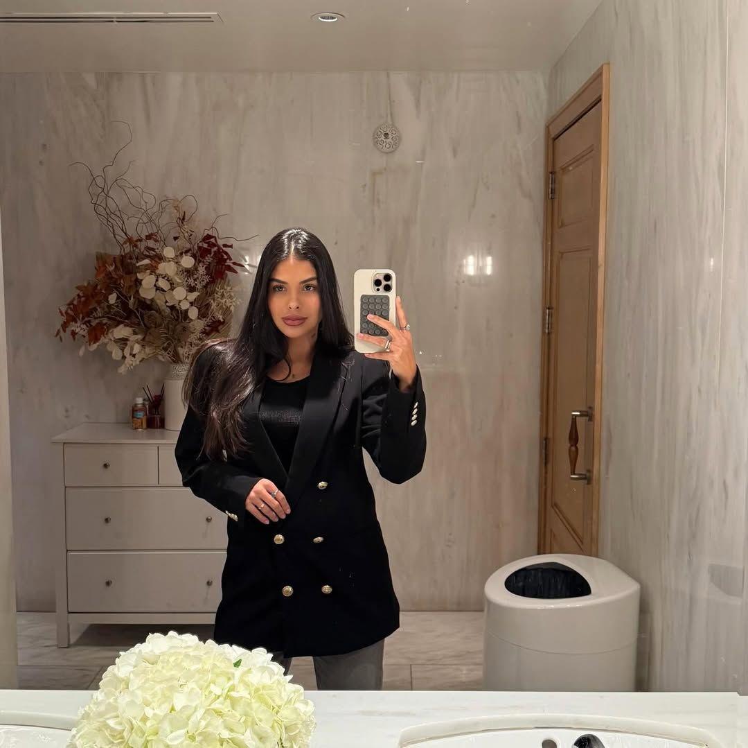 Munik Nunes vestindo blazer preto elegante tirando uma selfie no espelho em um banheiro ou closet com paredes de mármore.