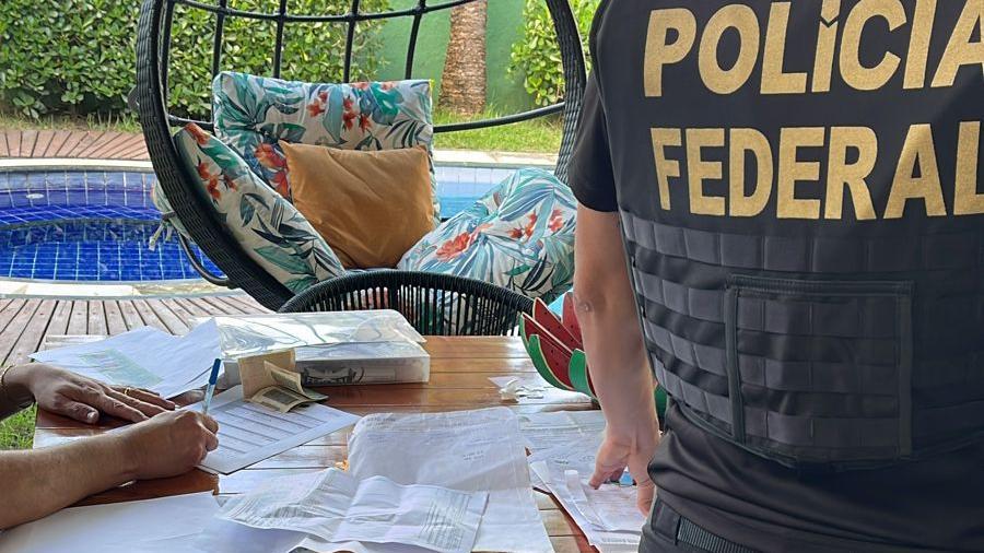 A imagem mostra policiais federais realizando busca por documentos, que estão em cima de uma mesa, em uma casa. Um policial federal está fardado, de costas.
