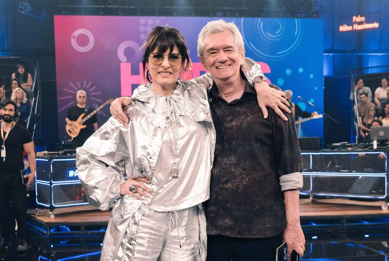 Homem e mulher brancos abraçados e sorrindo para a câmera. À esquerda, a mulher tem cabelo preto, liso e curto, com óculos escuros e roupa prateada. À direita, o homem tem cabelos grisalhos e roupa social preta.