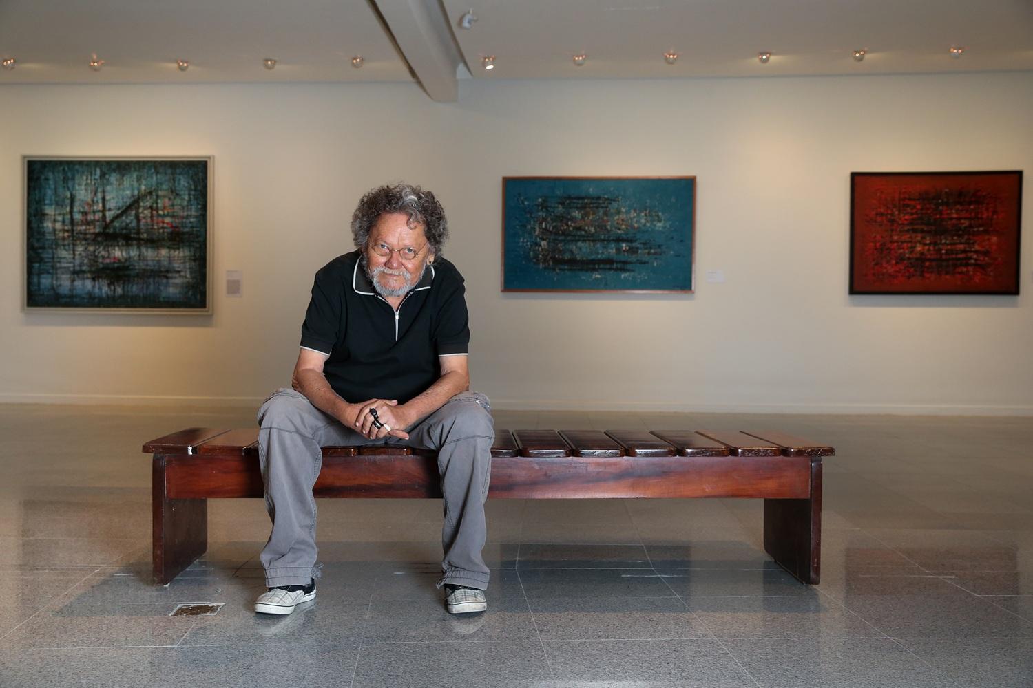 O artista Zé Tarcísio está sentado em um banco de madeira no centro de uma galeria de arte. Ao fundo, três quadros abstratos estão pendurados em uma parede branca iluminada.